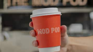Eine Hand hält eine rote Kaffeetasse mit der Aufschrift "MOD POD", vor einem Imbisswagen mit der Beschilderung "THE WOO POD".