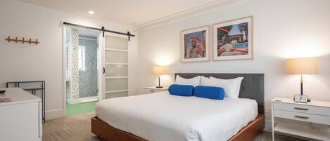 A modern hotel room with a large bed, two bedside tables with lamps, a wardrobe, and a sliding glass door to the bathroom.