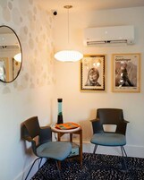A room with two chairs, a small table, a wall-mounted air conditioner, and two framed pictures.
