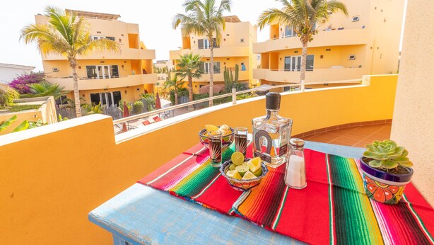 A balcony with a table set with drinks and a salt shaker, overlooking yellow buildings and palm trees.