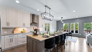 A modern kitchen with light wood cabinets, a central island with bar stools, and a large window offering a view of greenery. - Elysian Shores W/heated Pool & Optional Boat Rental (Mooresville)