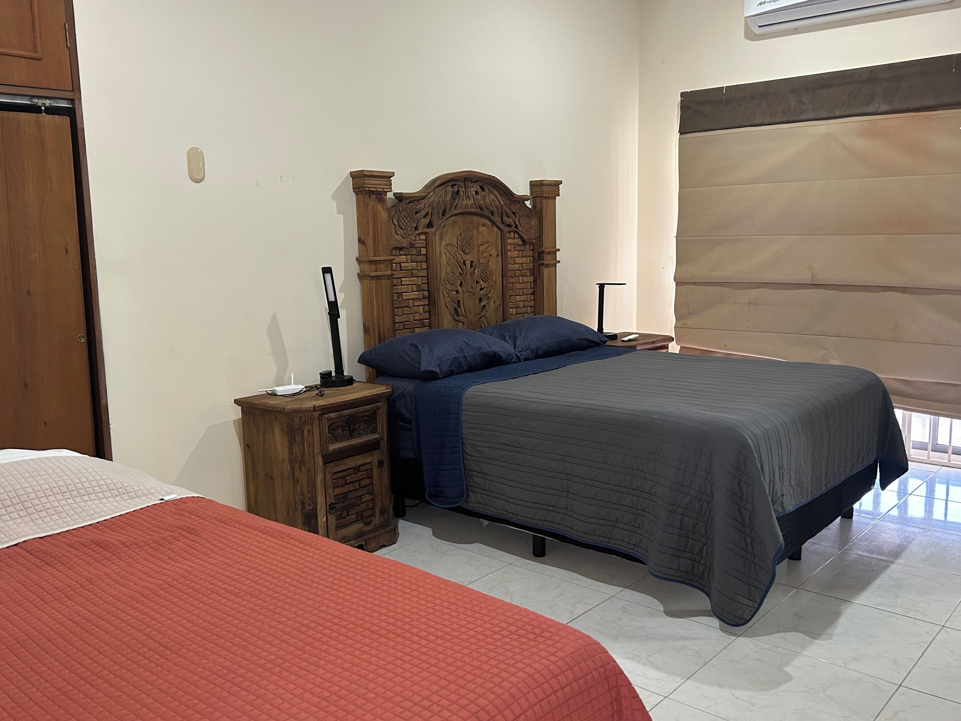 Un dormitorio con cabecera de madera, mesita de noche con lámpara, cama con colcha azul y gris, y ventana con persiana.