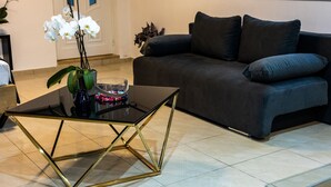 A modern living room with a dark grey sofa, a glass coffee table with gold legs, and a vase with white orchids. - Black suites 1 (Chania)