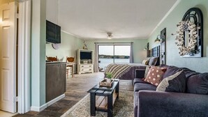 Un salon avec un canapé de couleur sombre, une table basse et un téléviseur fixé au mur. Une fenêtre offre une vue sur l’extérieur.