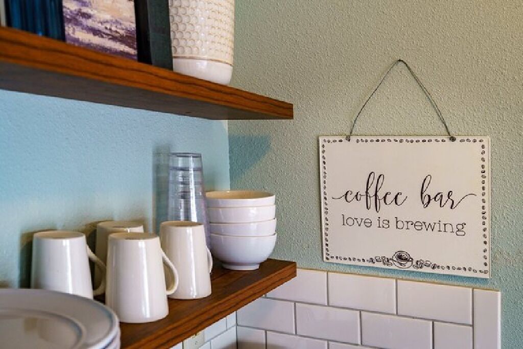 A kitchen area with white mugs, bowls, and a coffee maker on a shelf. A sign on the wall reads "coffee bar love is brewing."