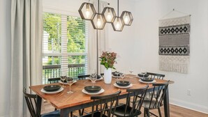 Una sala da pranzo con un tavolo di legno apparecchiato per sei persone, sedie nere, un lampadario esagonale sospeso e un arazzo appeso al muro.