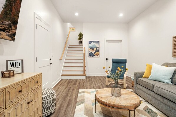 Un salon moderne avec un escalier, un canapé gris agrémenté de coussins jaunes et bleus, une table basse en bois sur laquelle est posé un vase de fleurs, et un cadre photo fixé au mur.