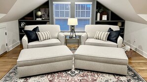 A loft-style room with two grey armchairs, a small side table, and a floor lamp. There are two windows with blinds, a bookshelf, and a decorative globe. - Lofty Dream | Stylish Northshore Loft Stay (Chattanooga)