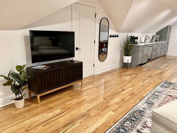 A modern living room with a wooden floor, a dark wooden entertainment unit, a flat-screen TV, a large mirror, and a kitchen area with cabinets and a countertop.
