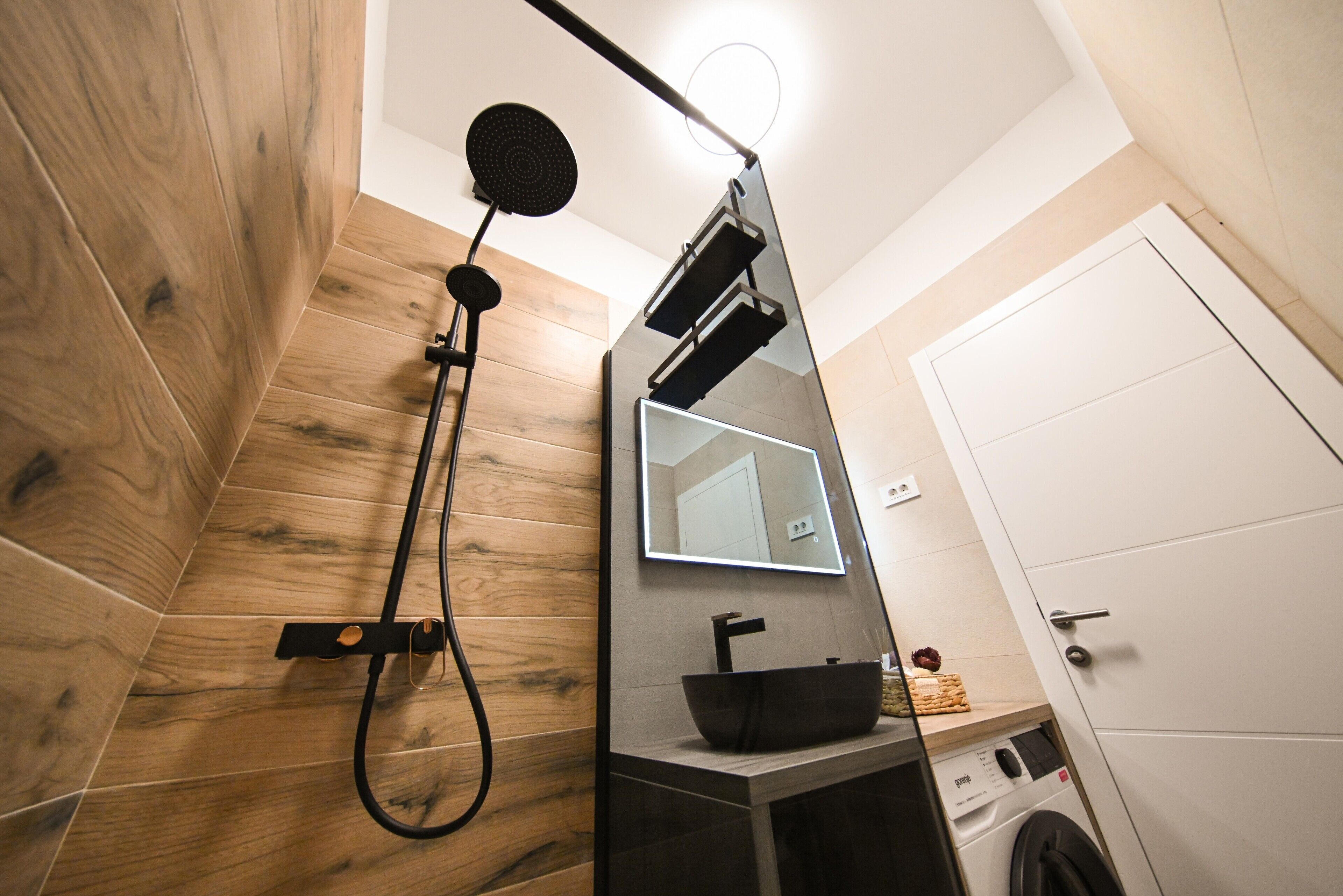 Ein modernes Badezimmer mit Holzboden, Waschbecken in Schwarz, einem Spiegel und einer Dusche mit Duschbrause.