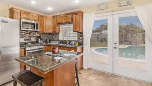 A kitchen with wooden cabinets, stainless steel appliances, a granite countertop, and a view of a swimming pool through glass doors. - Quaint Coastal Pool Home Oasis Florida Space Coast, by Beach&Space Coast Stadium (Melbourne)