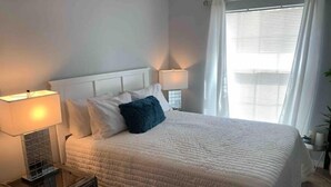A neatly made bed with a white duvet and pillows, a bedside table with a lamp, and a window with curtains. - Quaint Coastal Pool Home Oasis Florida Space Coast, by Beach&Space Coast Stadium (Melbourne)