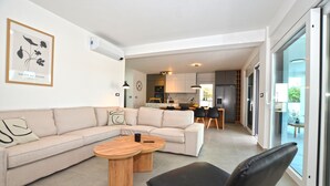 A modern living room with a beige sectional sofa, two wooden coffee tables, and a black chair. A kitchen is visible in the background with modern appliances and a dining area. - Villa 5H (Kanfanar)