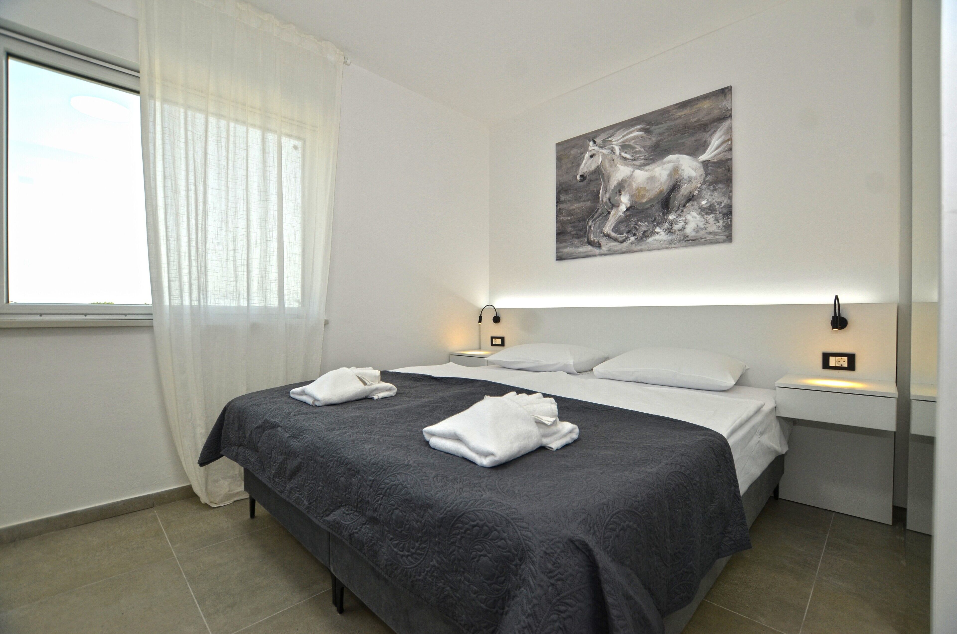 A neatly made bed with a dark patterned bedspread, white pillows, and a folded white towel on the bed. There is a painting of a horse on the wall above the bed.