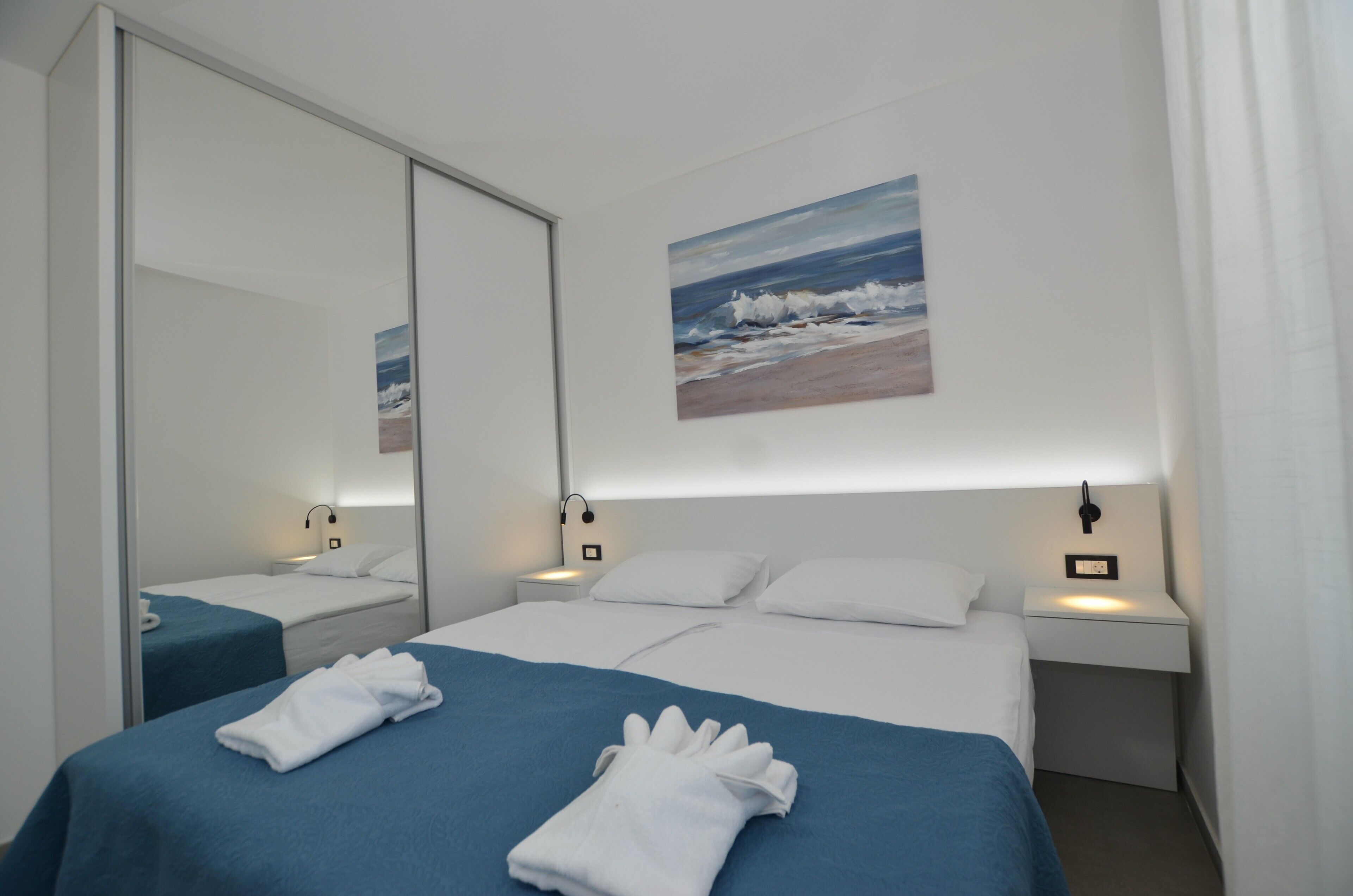 Una camera d'albergo con due letti, un grande specchio, un quadro raffigurante una spiaggia e delle lampade ai comodini.