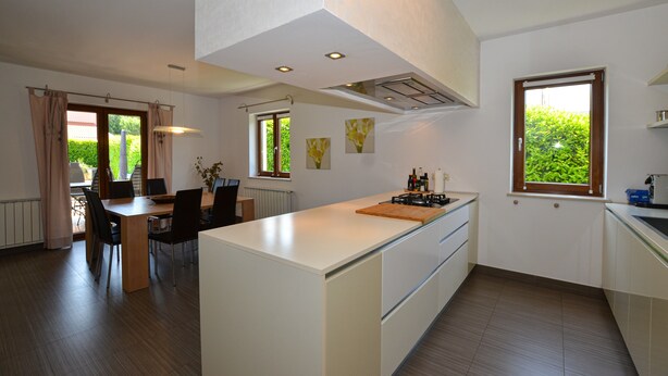 A modern kitchen with a central island, dining area with chairs, and large windows offering views of greenery. - Villa dado (Kanfanar)