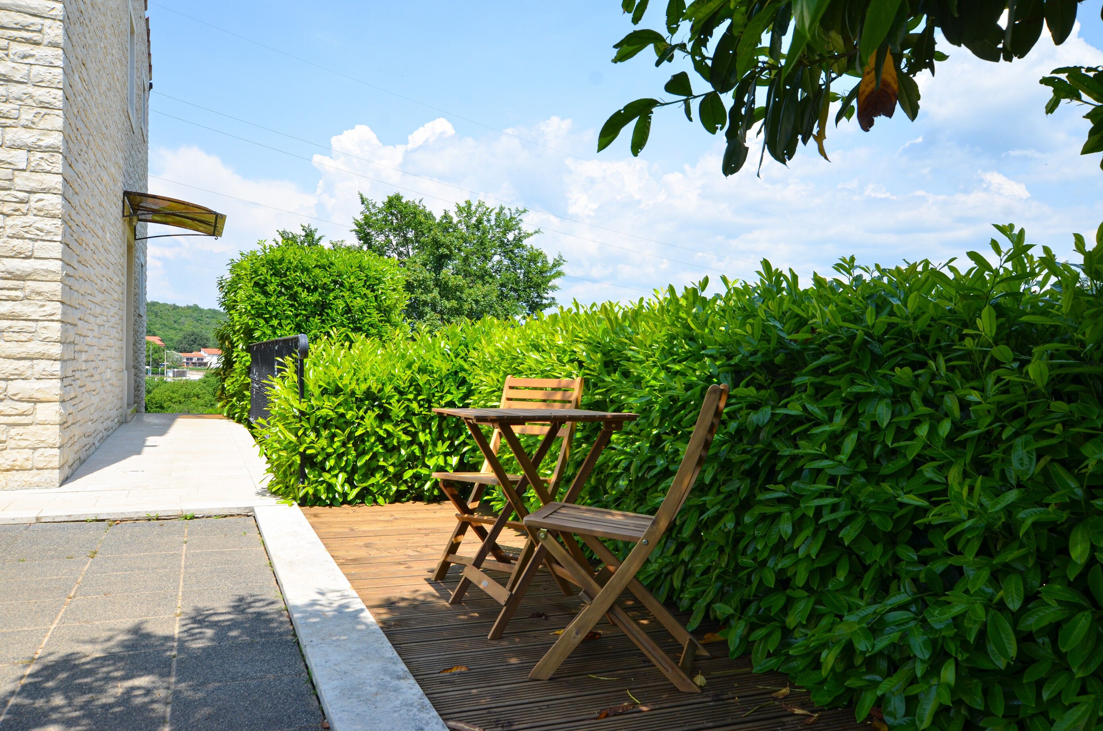 A wooden deck with a table and chairs, surrounded by lush greenery and a stone wall.