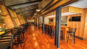 Un comedor alargado y estrecho con suelo de madera y sillas de metal negro. Se observan varias mesas y una zona de bar con taburetes.