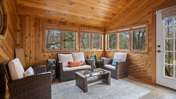 Interior acogedor de una cabaña con paredes de madera, ventanas amplias, muebles de mimbre y una mesa de centro de vidrio.