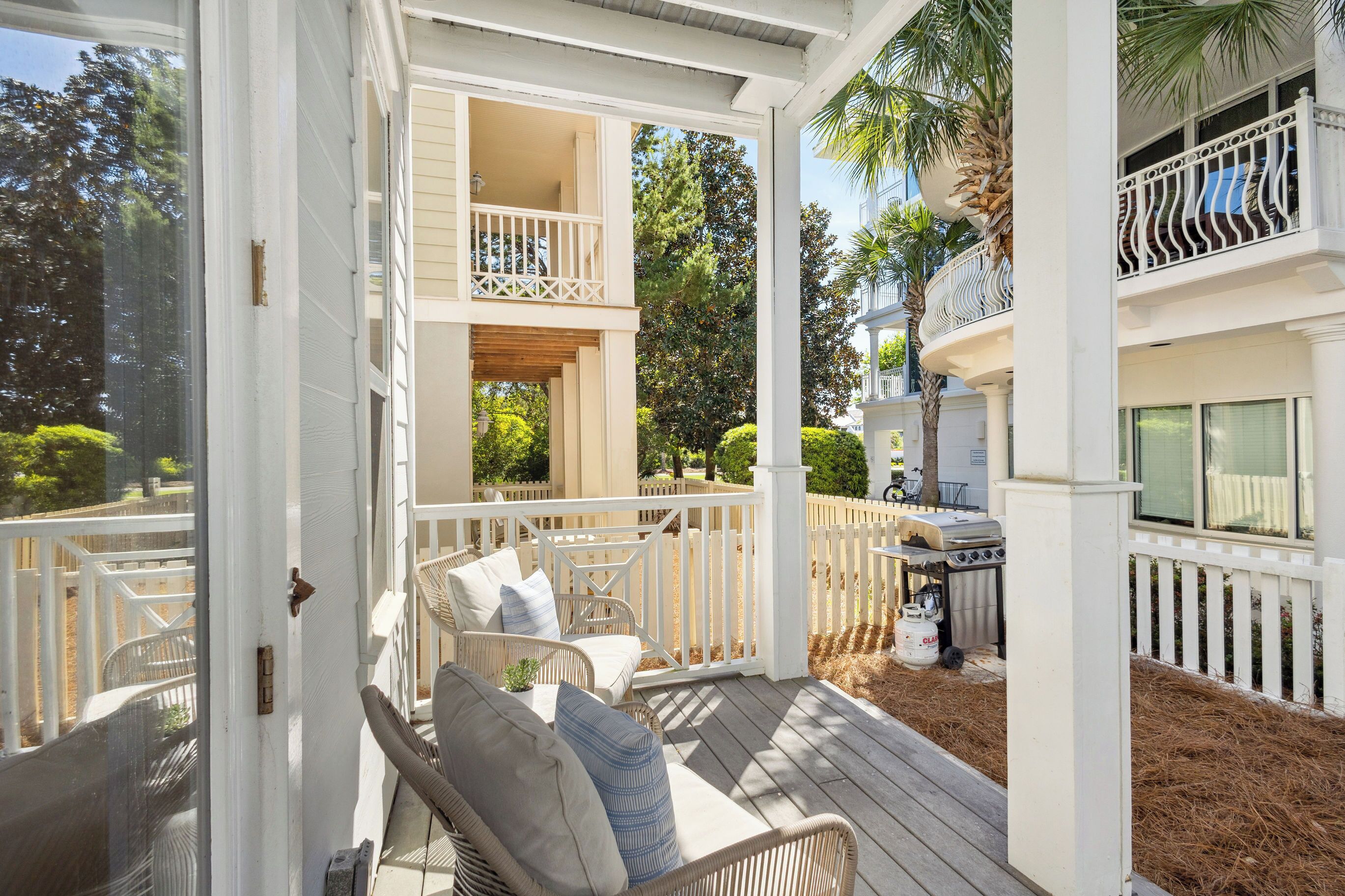 A covered outdoor deck with wicker furniture, a grill, and a view of a residential building with a balcony.