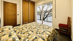 Un dormitorio con una cama grande, una puerta de madera, una puerta corredera de vidrio que da a un balcón con barandilla y árboles.