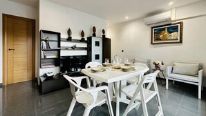 Un comedor moderno con una mesa blanca dispuesta para cuatro personas, rodeada de sillas blancas. Un estante con objetos decorativos está contra la pared, y un cuadro encima de la mesa del comedor.