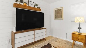 A room with a large flat-screen TV on a wooden TV stand, a small wooden nightstand with a lamp, and a patterned rug on the floor. - The Gator Getaway- Huge Backyard (Gainesville)