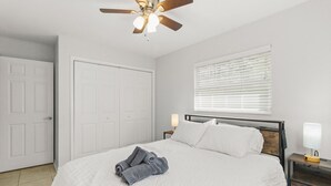 A bedroom with a bed, a ceiling fan, a window with blinds, a nightstand with a lamp, and a door. - The Gator Getaway- Huge Backyard (Gainesville)
