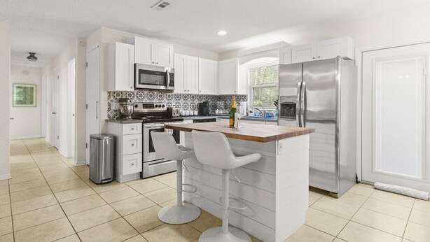 A modern kitchen with white cabinets, stainless steel appliances, a central island with bar stools, and a tiled backsplash. - The Gator Getaway- Huge Backyard (Gainesville)