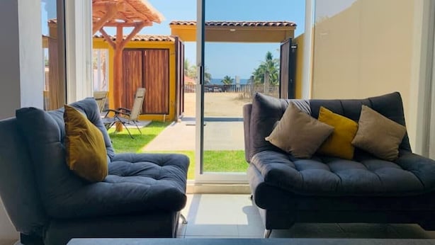 Una habitación con dos sofás oscuros, una mesa de madera y un gran ventanal corredero que da a un espacio exterior con terraza de madera y un paraguas amarillo.