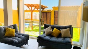 Una habitación con dos sofás de cuero negro y cojines amarillos, un ventanal y vistas a un patio con una pérgola de madera y una mesa.