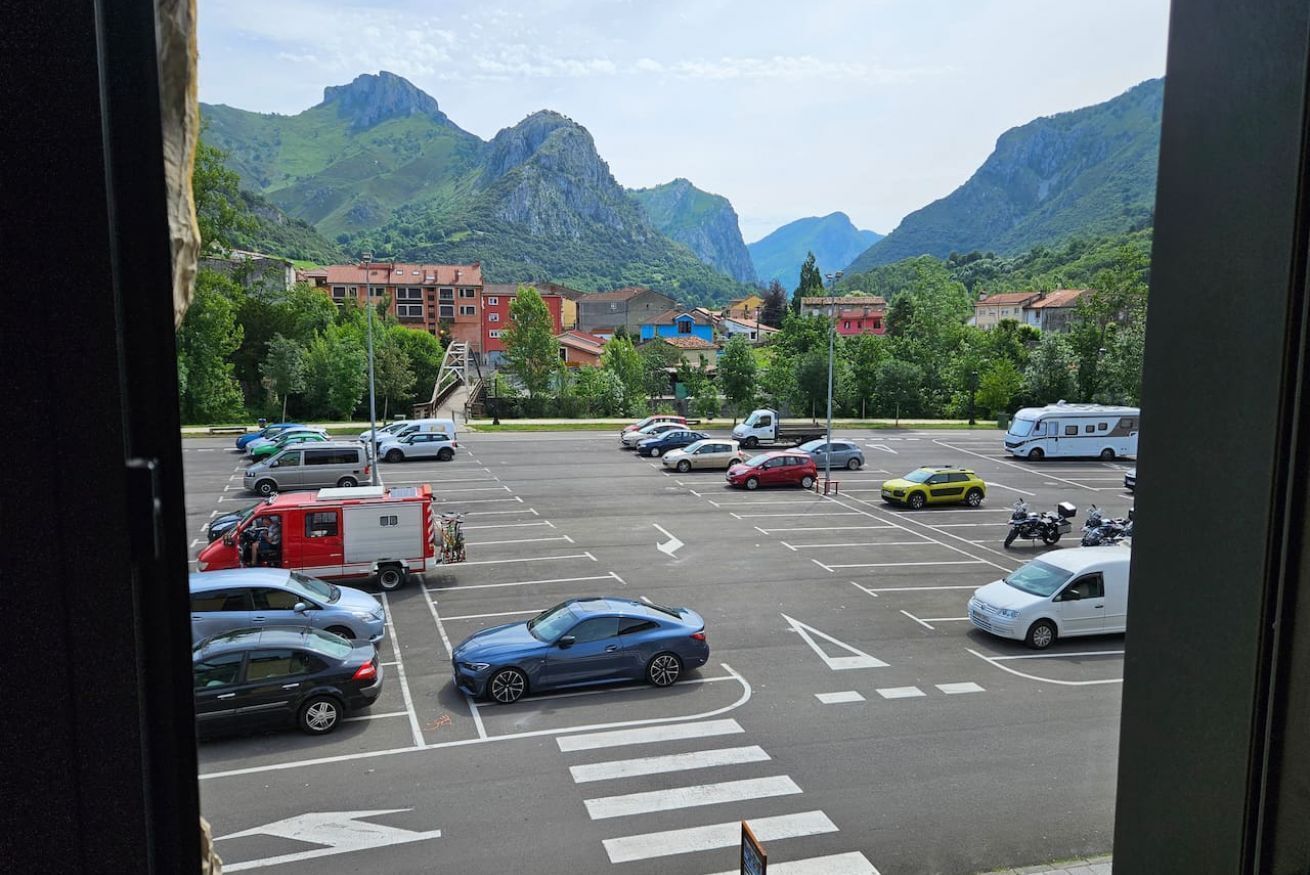 Un estacionamiento con coches y una caravana, rodeado de edificios y montañas.