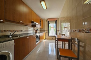 Cocina con armarios de madera, horno empotrado, lavadora y comedor con mesa y sillas.