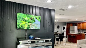 A modern living room with a flat-screen TV mounted on a dark wall, a glass cabinet below, and a dining area with a table and chairs. - Unique space with lots of details! (Seffner)