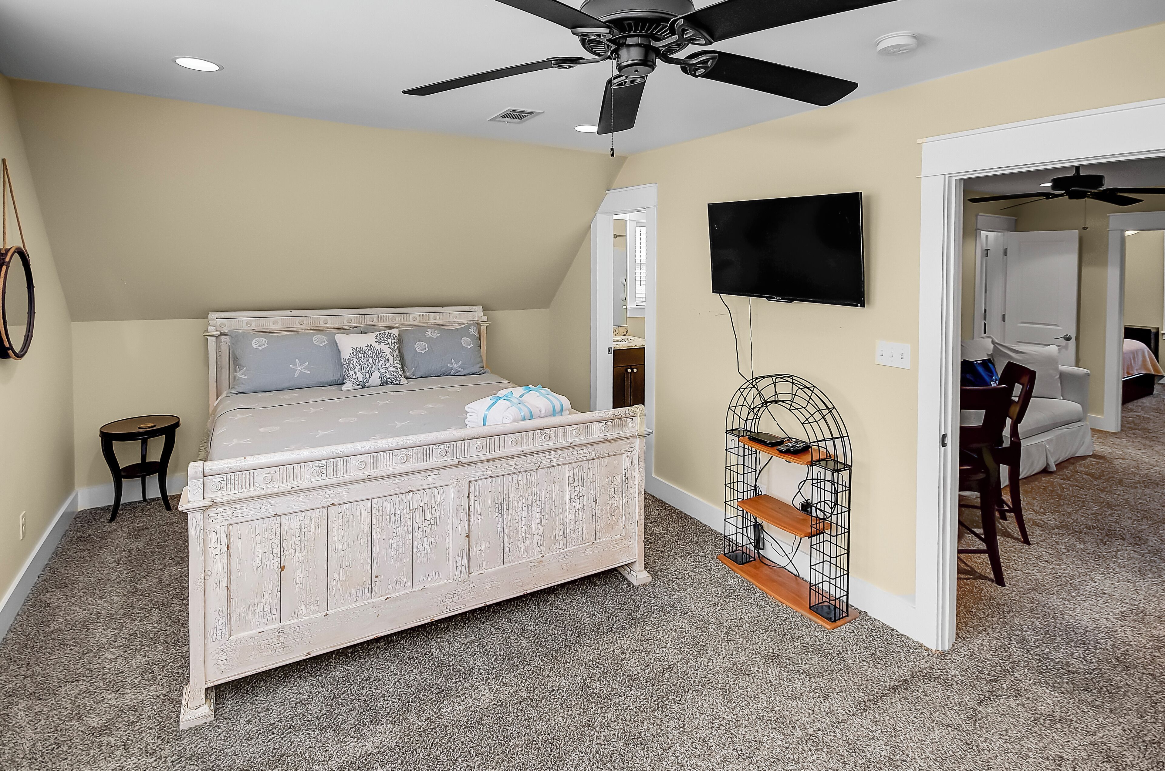 A bedroom with a white bed, a ceiling fan, a TV mounted on the wall, and a small side table.