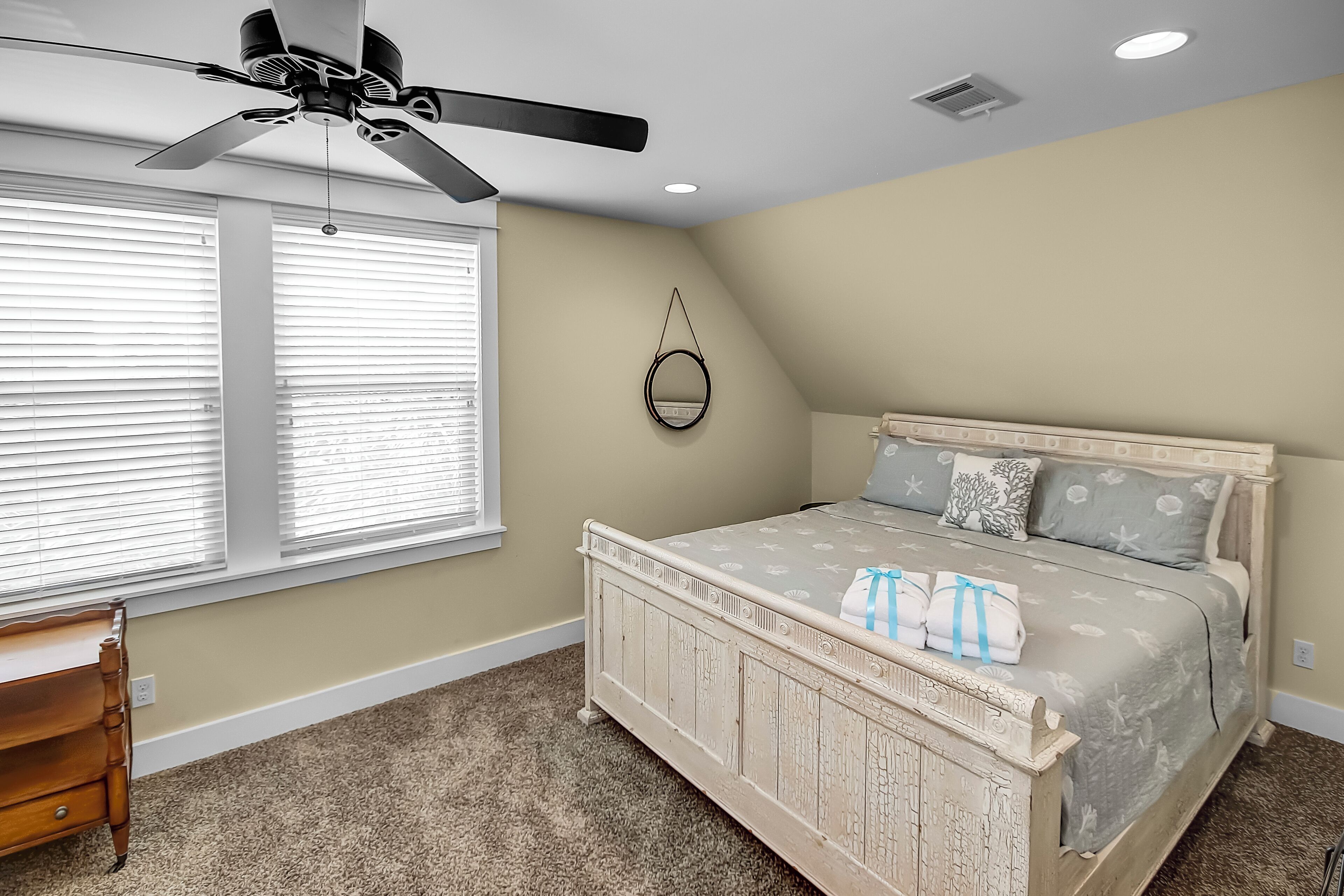 A bedroom with a bed, a ceiling fan, a window with blinds, and a small wooden dresser.