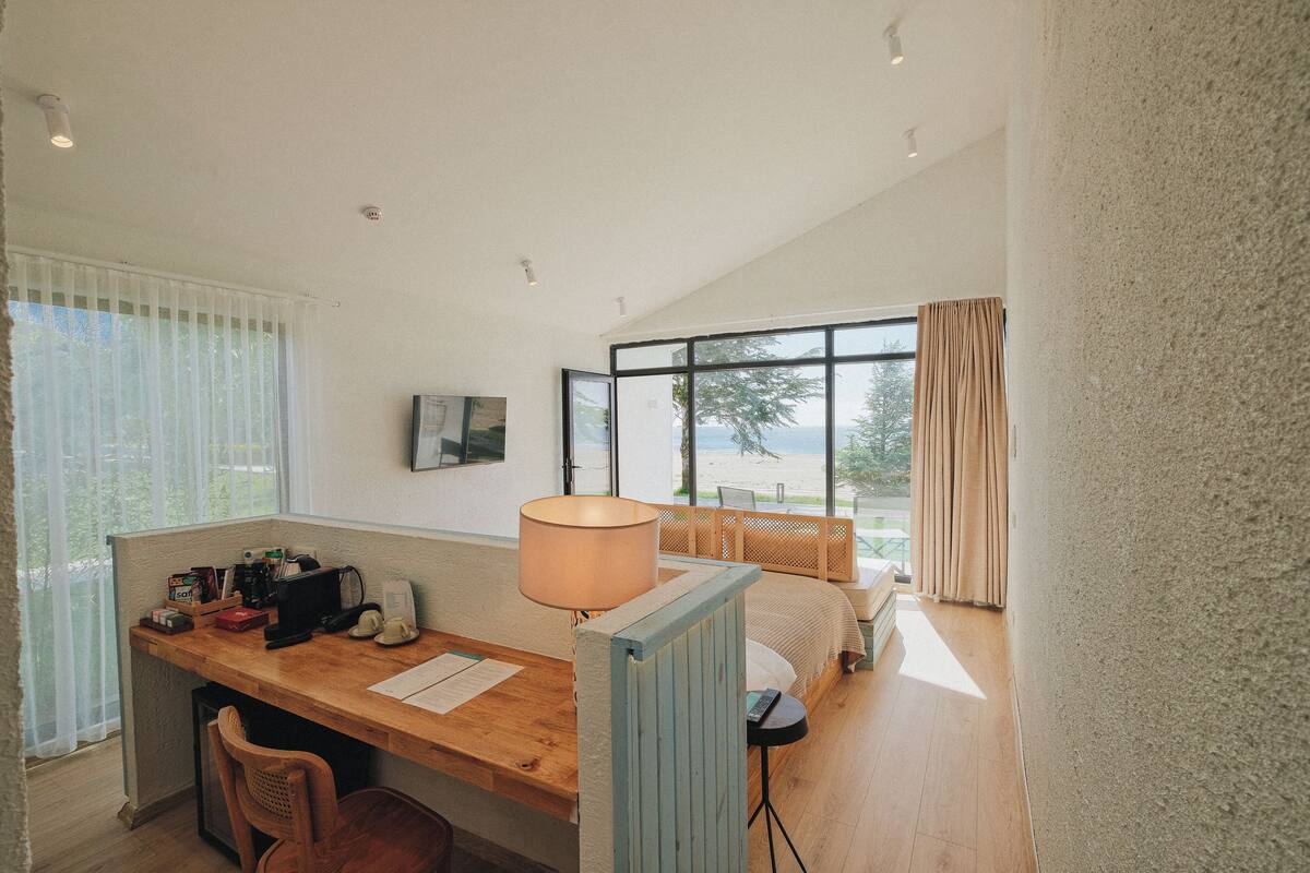 A room with a wooden desk, a chair, a bed with a view of the beach, and a large window with curtains.