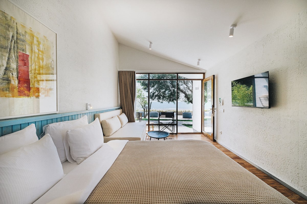 A modern hotel room with a large bed, a sofa, a flat-screen TV, and a view of the outdoors.