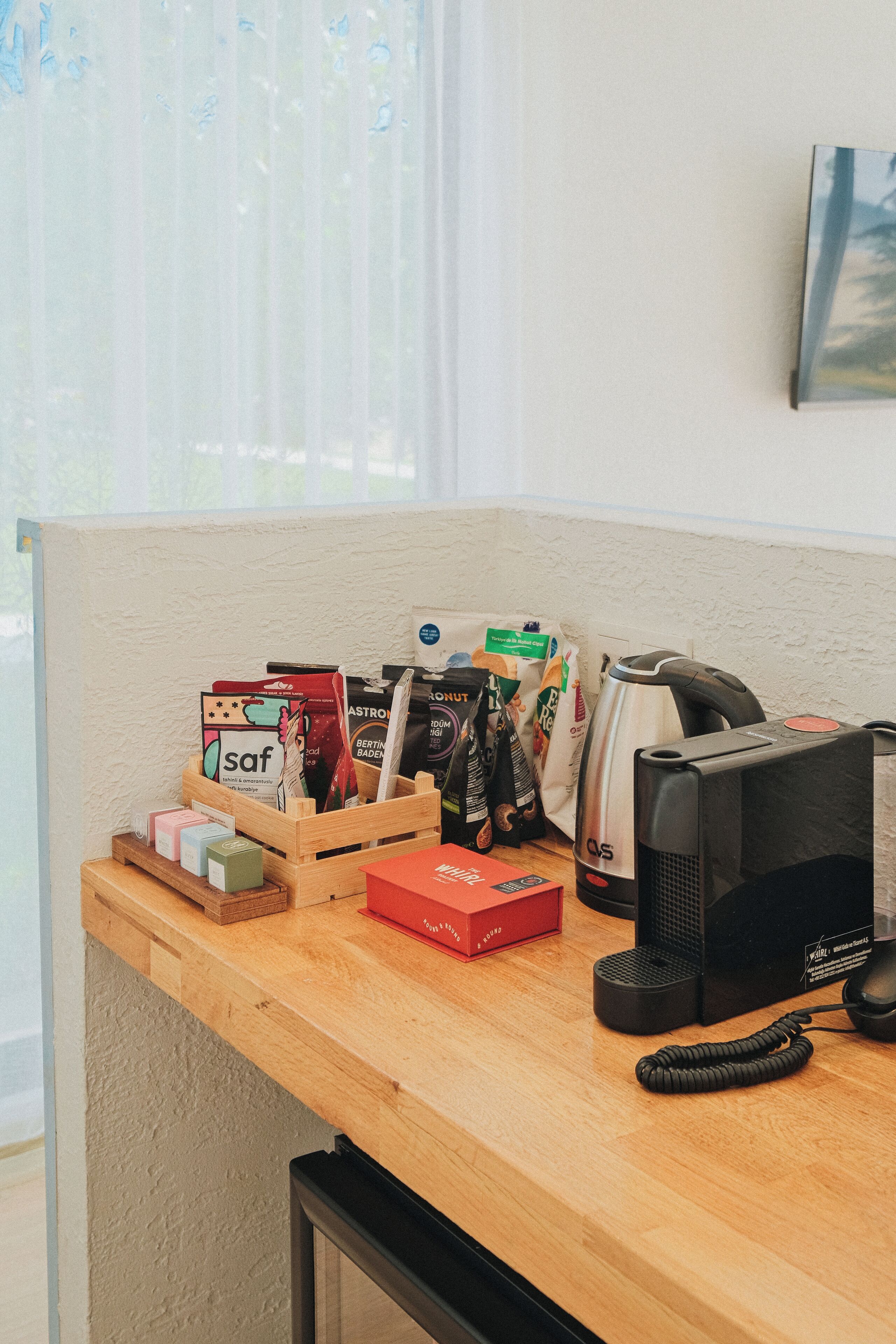 Un escritorio de madera con una cafetera, una computadora y varios objetos, incluyendo una caja roja y un porta tarjetas con cápsulas de café.