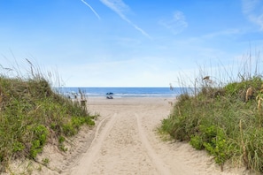 Un sentier de sable qui longe la plage et mène à l’océan, sous un ciel dégagé.