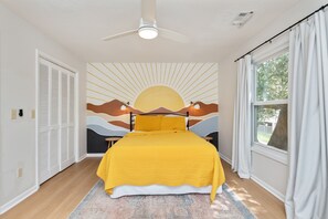 Une chambre à coucher avec un lit, un ventilateur de plafond, une fenêtre avec des rideaux et une fresque murale.