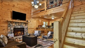 L’intérieur d’une cabane en rondins est agrémenté d’une cheminée en pierre, d’un téléviseur à écran plat fixé au-dessus, d’un coin salon confortable avec une table basse et d’un escalier menant à un étage supérieur.