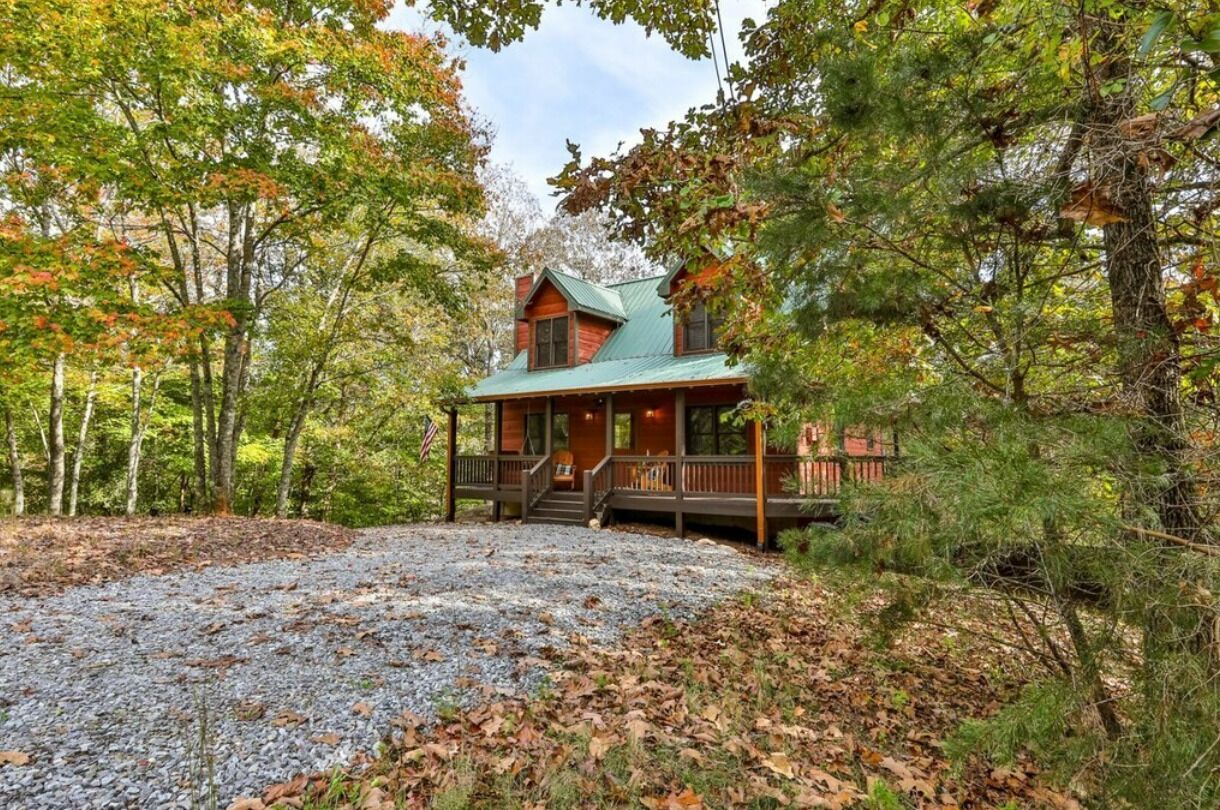 A cabin with a porch, surrounded by trees and a gravel driveway.