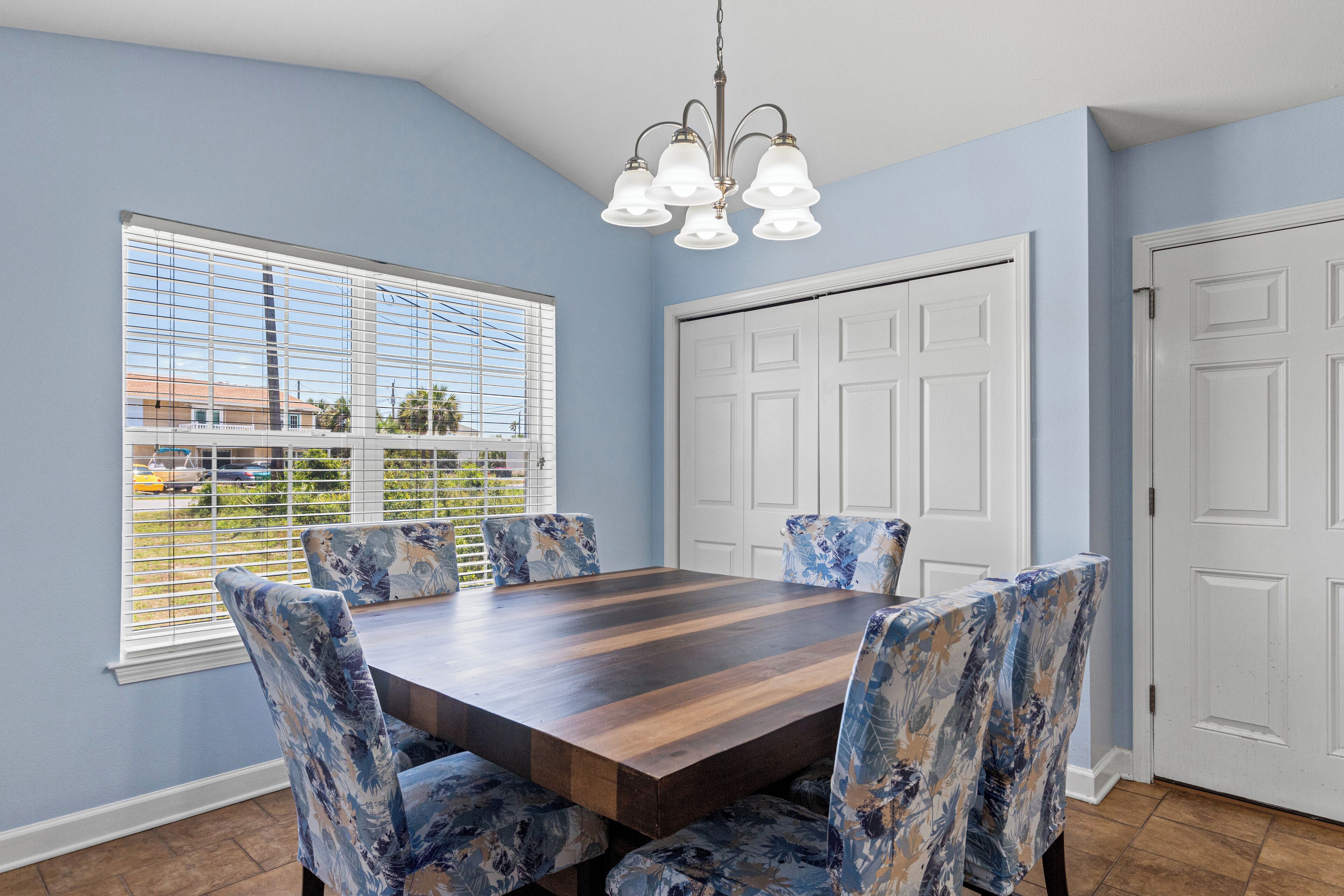 Un comedor con una mesa de madera rectangular rodeada de sillas con estampado floral, una araña y un gran ventanal con persianas.