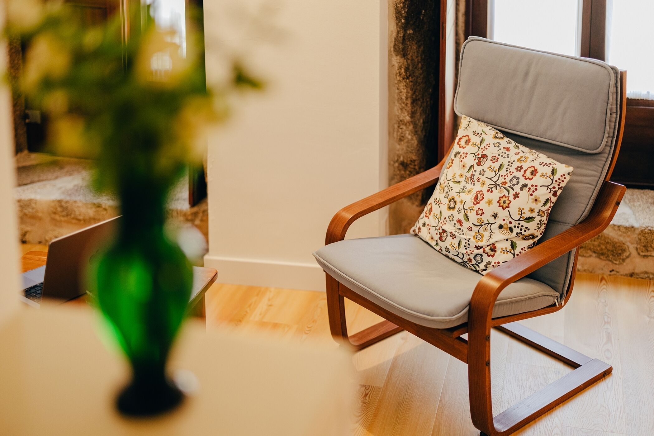 A wooden chair with a cushion and a floral pillow, positioned near a window with a view of a cityscape.