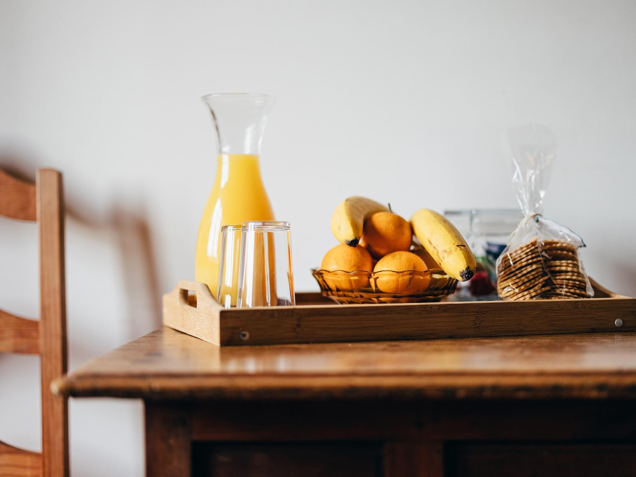 Una mesa de madera con una bandeja que contiene una jarra de jugo de naranja, dos vasos, una cesta de naranjas, plátanos y una hogaza de pan.