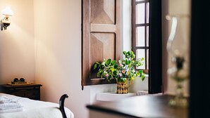 Et soveværelse med en seng, et natbord, en lampe, et vindue med en trælugt og en vase med blomster på vindueskarmen.
