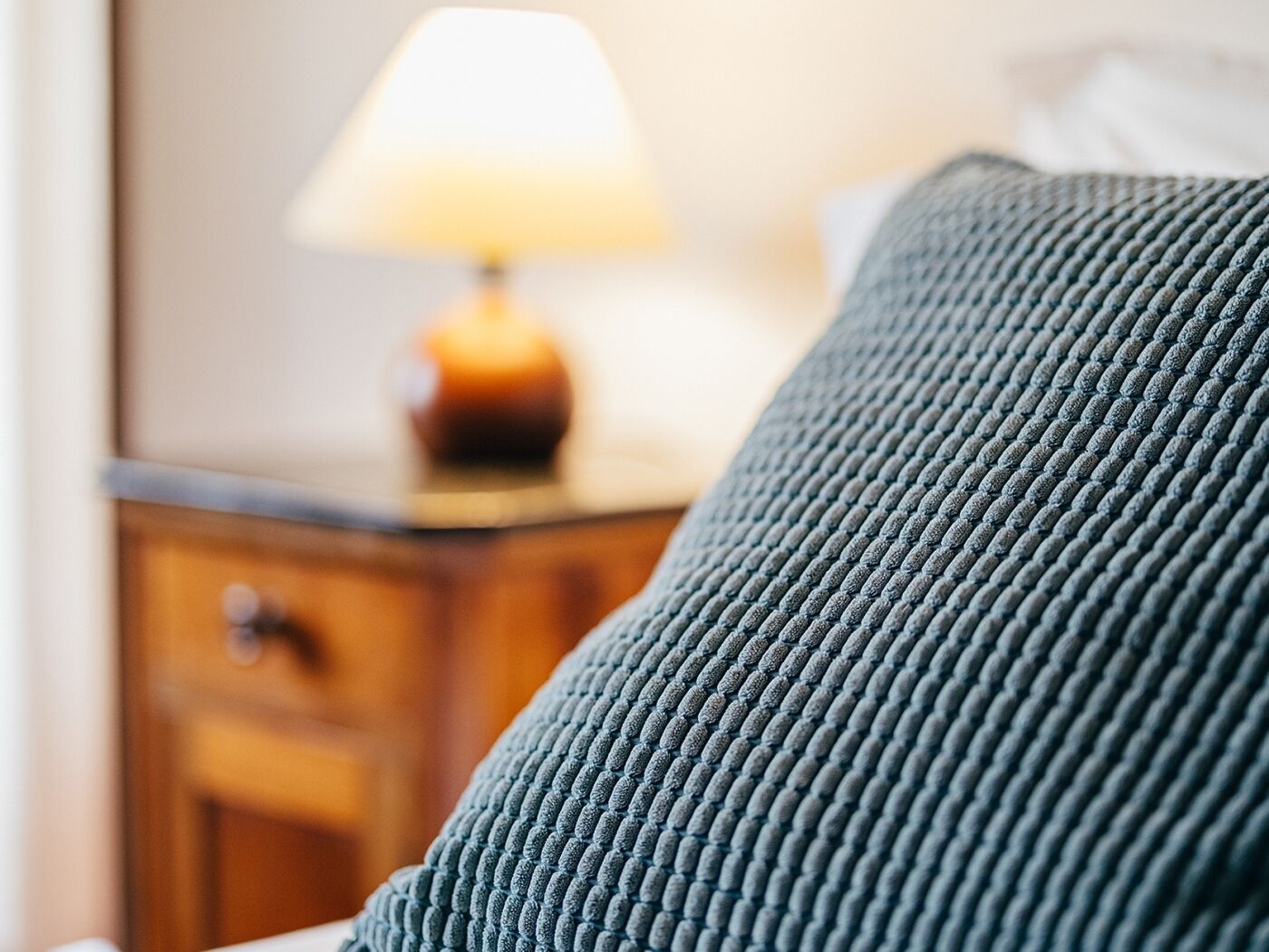 Primer plano de una tela azul con textura, probablemente un sofá o cojín, sobre un fondo difuso que muestra una lámpara y muebles de madera.