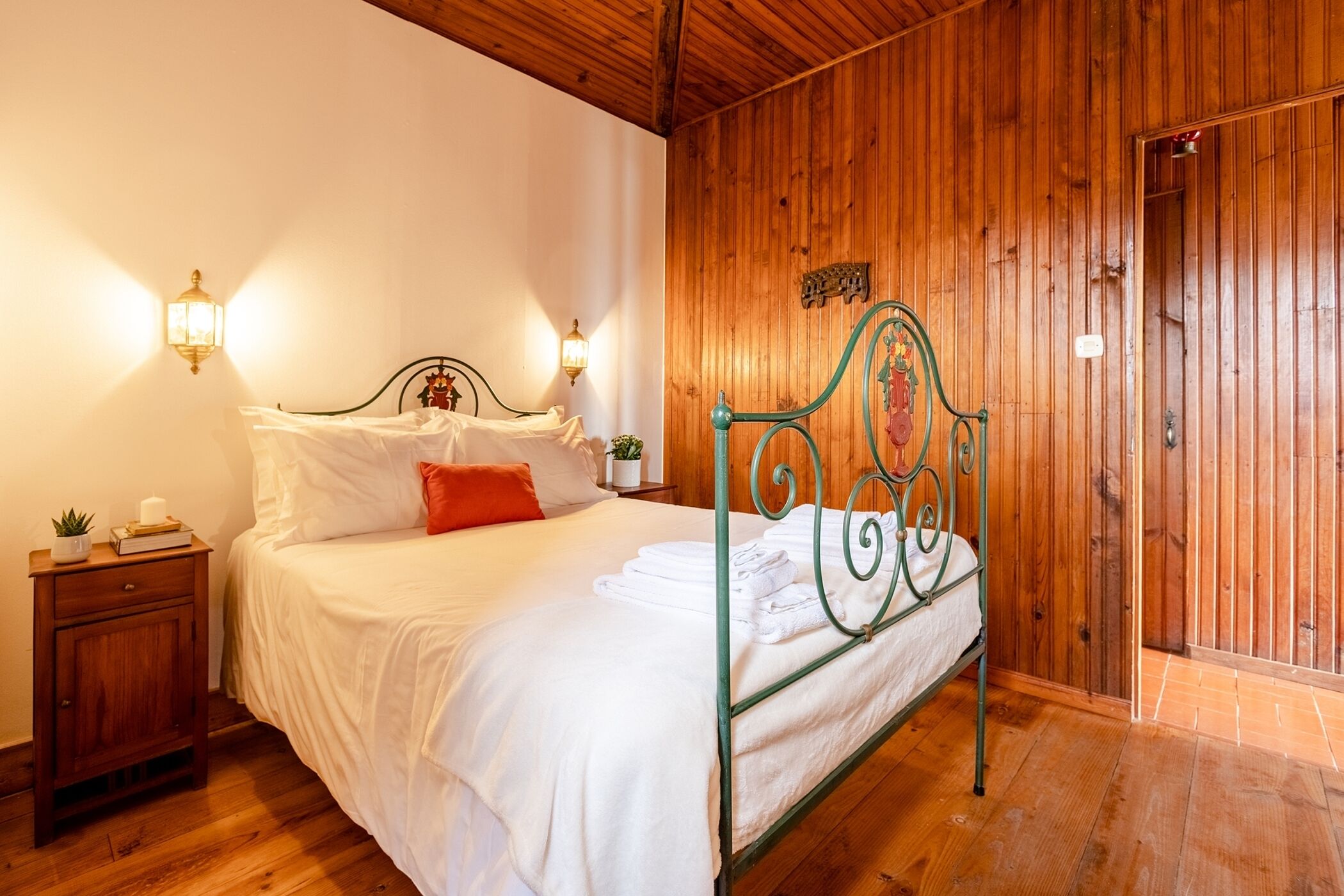 Una habitación con paneles de madera, una estructura de cama metálica, ropa de cama blanca y dos lámparas fijadas a la pared.