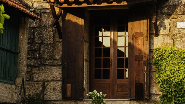 A stone building with a wooden door and shutters, a tiled roof, and a table with chairs outside.